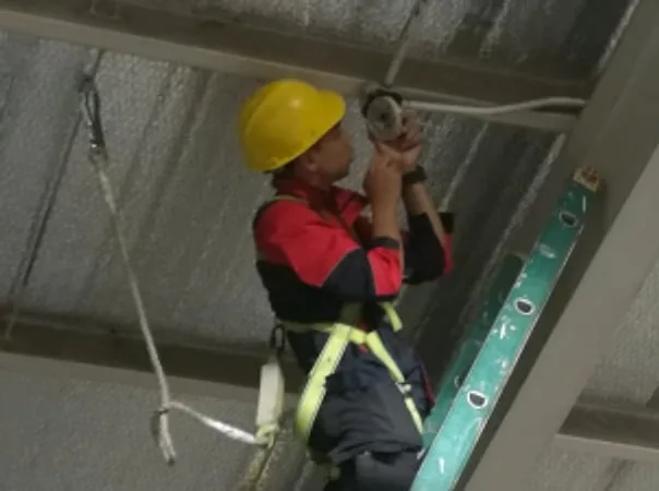 Technician in a hard hat and safety harness on a ladder installing a ceiling-mounted device