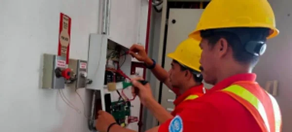 Two technicians in hard hats and safety vests testing a wall-mounted control box
