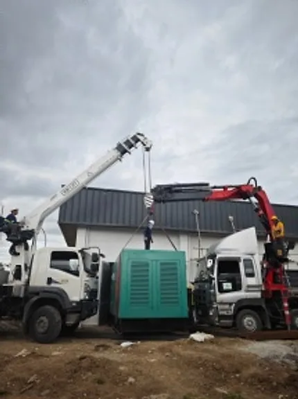 Two boom trucks lifting a green generator set on a work site