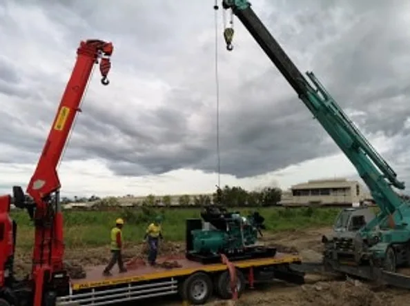 Two cranes lifting a generator set onto a flatbed truck in an open field